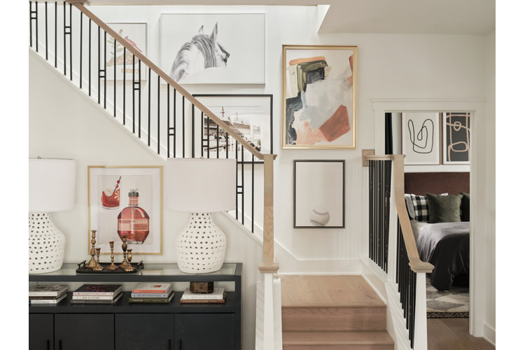 A stylish home entryway positioned next to a wooden staircase with black metal balusters. On the left, a black console table with lower shelving holds books, a tray with brass candlesticks, and two large white textured ceramic table lamps with white drum shades