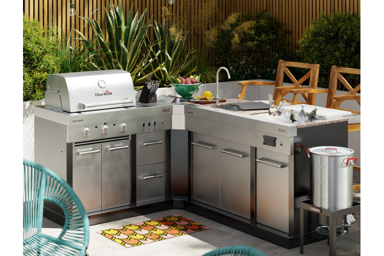 outdoor kitchen with an stainless steel outdoor sink, drink chiller, and a grill in front of a plant border and wooden fencing.