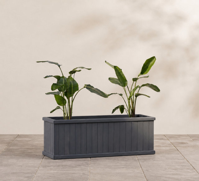Two green leafy plants potted in a rectangular charcoal grey slatted wooden planter set on a stone tiled patio.
