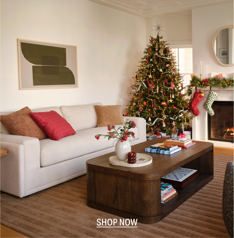 Cozy living room decorated for the holidays with a lit Christmas tree, a fireplace with stockings, and a light-colored sofa, featuring a coffee table with books and decor, Shop Now.