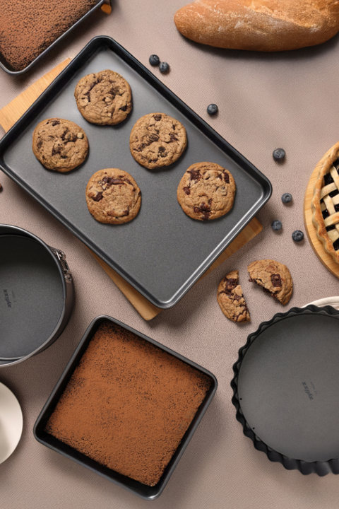 A variety of Zyliss non-stick bakeware including a rectangular baking sheet with chocolate chip cookies, a square pan with cocoa powder, a springform pan, and a tart pan, displayed on a table with a loaf of bread and a blueberry pie.