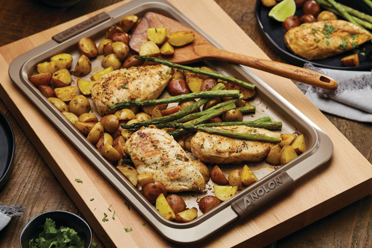 a silver sheet pan with chicken breasts potatoes and asparagus with a wooden spatula and a wood cutting board