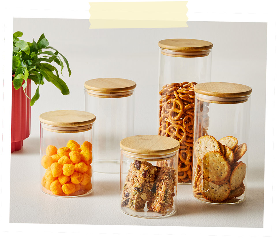Cinq bocaux de rangement en verre transparent avec couvercles en bambou remplis de diverses collations comme des bretzels, des craquelins et des grignotines.