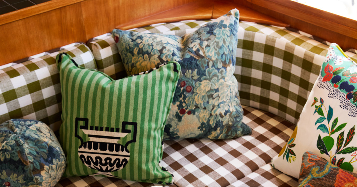 A corner seating area featuring brown and white checkered upholstery, accented with a variety of decorative throw pillows in mixed patterns including green stripes with a vase motif, floral tapestries, and a botanical print.