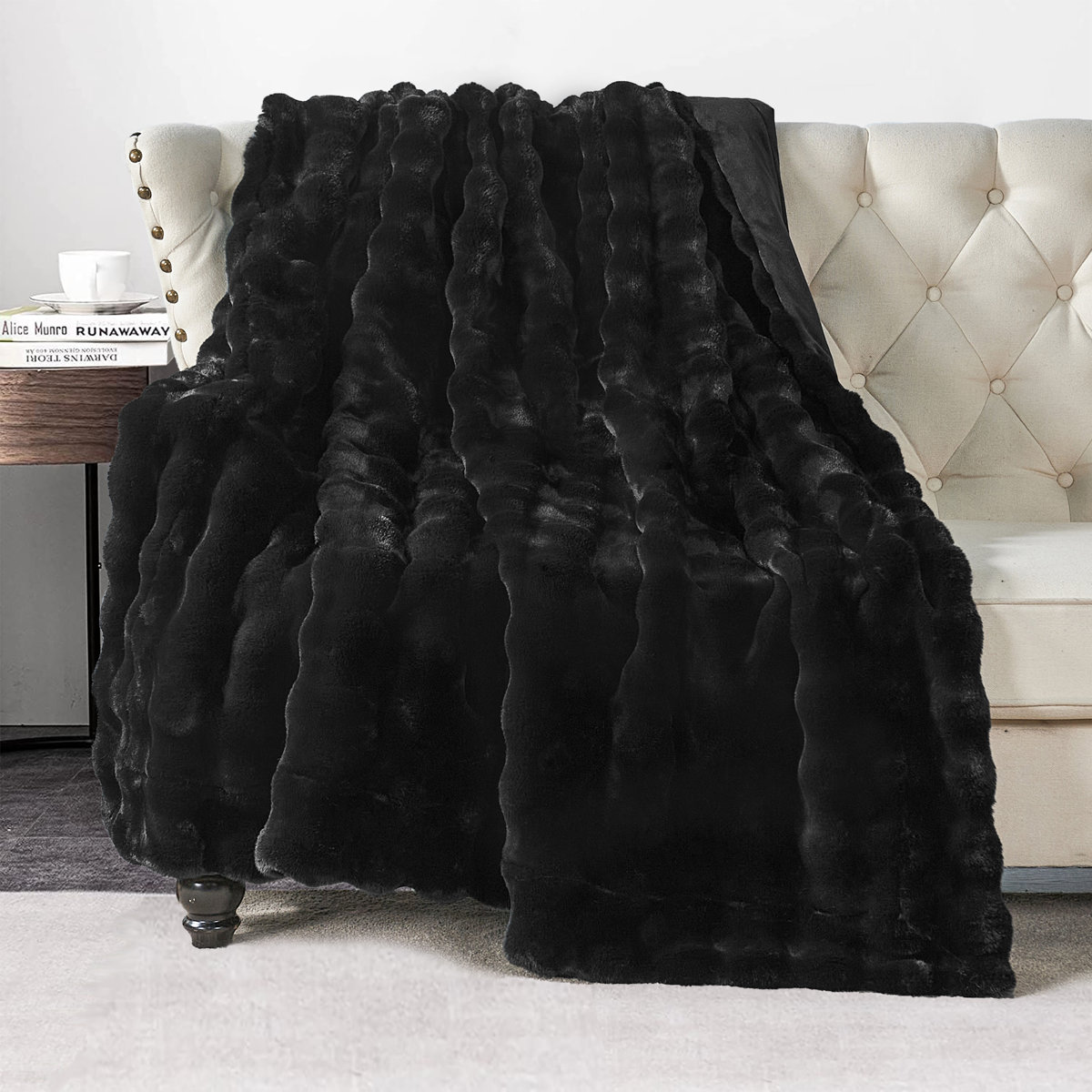 Soft black faux fur throw blanket draped over a tufted cream sofa beside a wooden table with books and a teacup