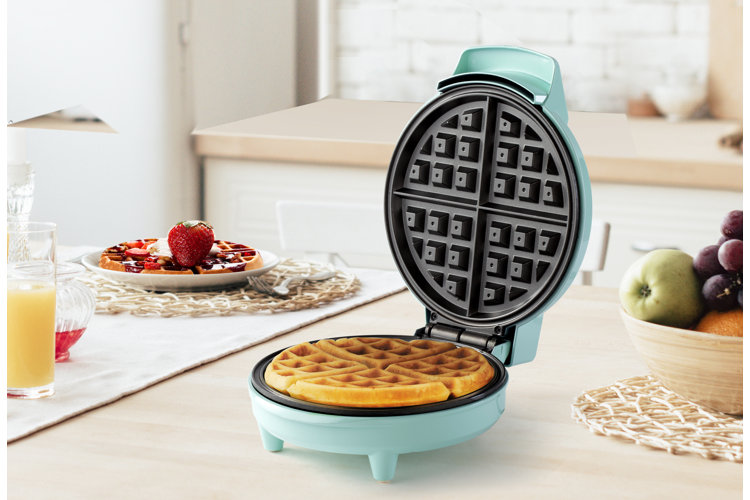 teal waffle maker on a wooden countertop next to a plate of waffles with chocolate syrup and strawberries.