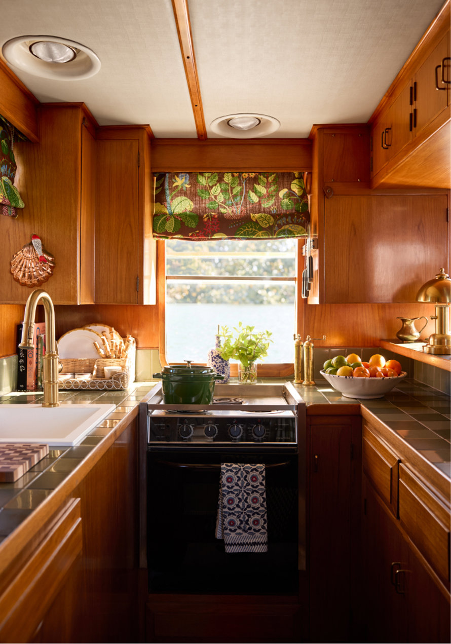 Cozy, well-lit kitchen on a boat featuring warm wood cabinetry, a centered stove with a green pot, a window overlooking water, and a marble countertop with fresh citrus and a brass faucet.