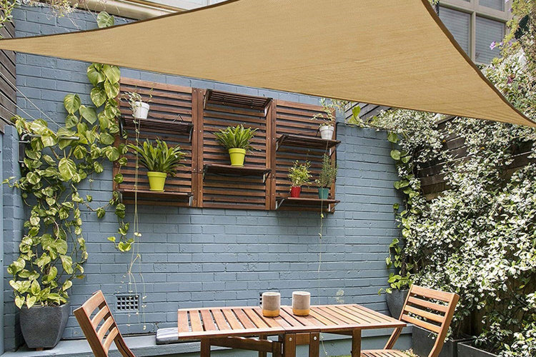 Shade sail idea with a beige triangular shade sail above a wood bistro set and surrounded by a brick wall and plants.