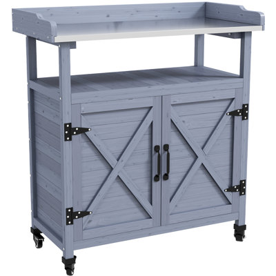 Table de rempotage grise en bois avec rangement intégré. Plateau en aluminium. Roulettes en caoutchouc pour un déplacement facile. Résistante aux intempéries. Parfait pour le jardinage. Dimensions : 88,3 cm (l) x 40,6 cm (p) x 95,2 cm (h). Table extérieure avec espace de rangement. Transport facile.
