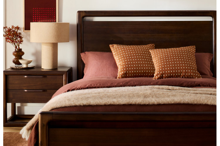 A cozy bedroom scene featuring a dark wood bed frame dressed in warm terracotta bedding, patterned rust-orange throw pillows, and a soft beige knit blanket draped across the foot.
