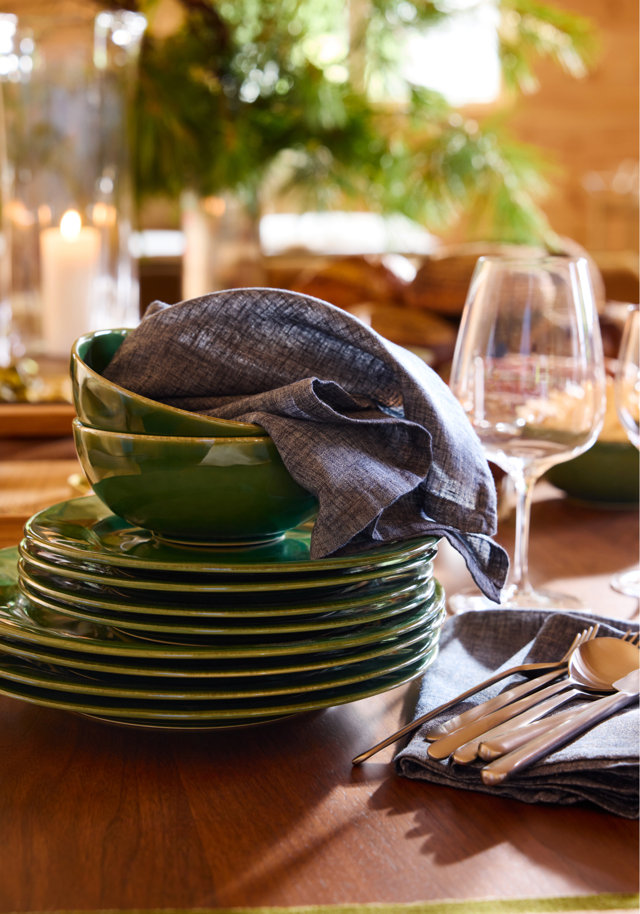 A stack of dark green ceramic plates and bowls topped with a blue linen napkin, placed next to a wine glass and polished silverware on a wooden dining table.