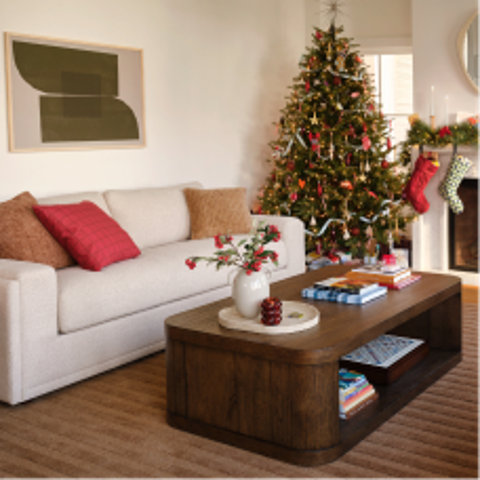 Living room decorated for the holidays with a lit Christmas tree, a beige sofa with red and brown throw pillows, and a dark wood coffee table with a white vase.