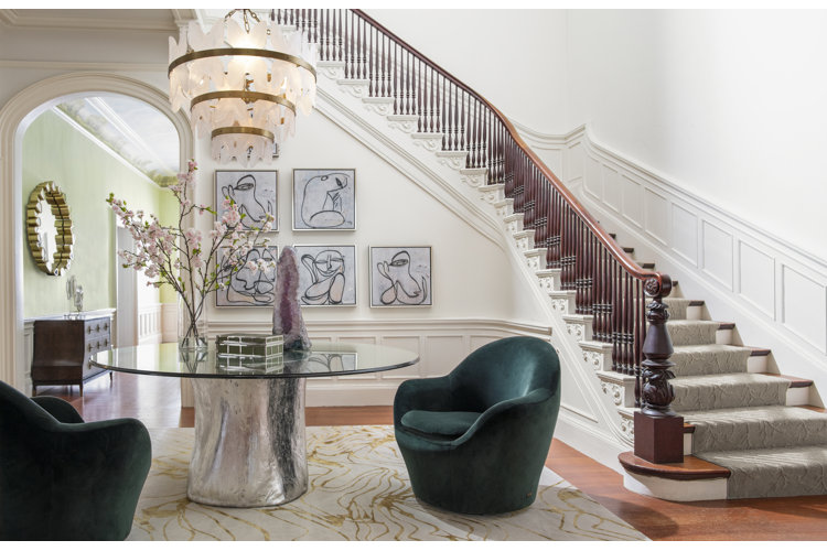 A large foyer with a curved staircase, a tiered chandelier, a modern pedestal table, two barrel chairs, and a gallery wall.