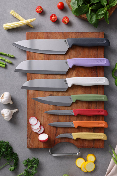 A set of seven colorful Zyliss kitchen knives arranged on a wooden cutting board, surrounded by fresh ingredients like cherry tomatoes, baby corn, garlic, asparagus, parsley, and radish slices.