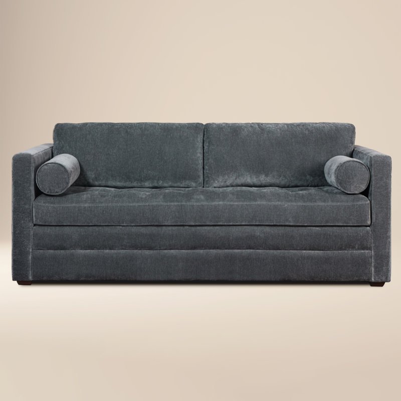 Dark grey velvet sofa with square arms, a tufted bench seat, and two cylindrical bolster pillows.