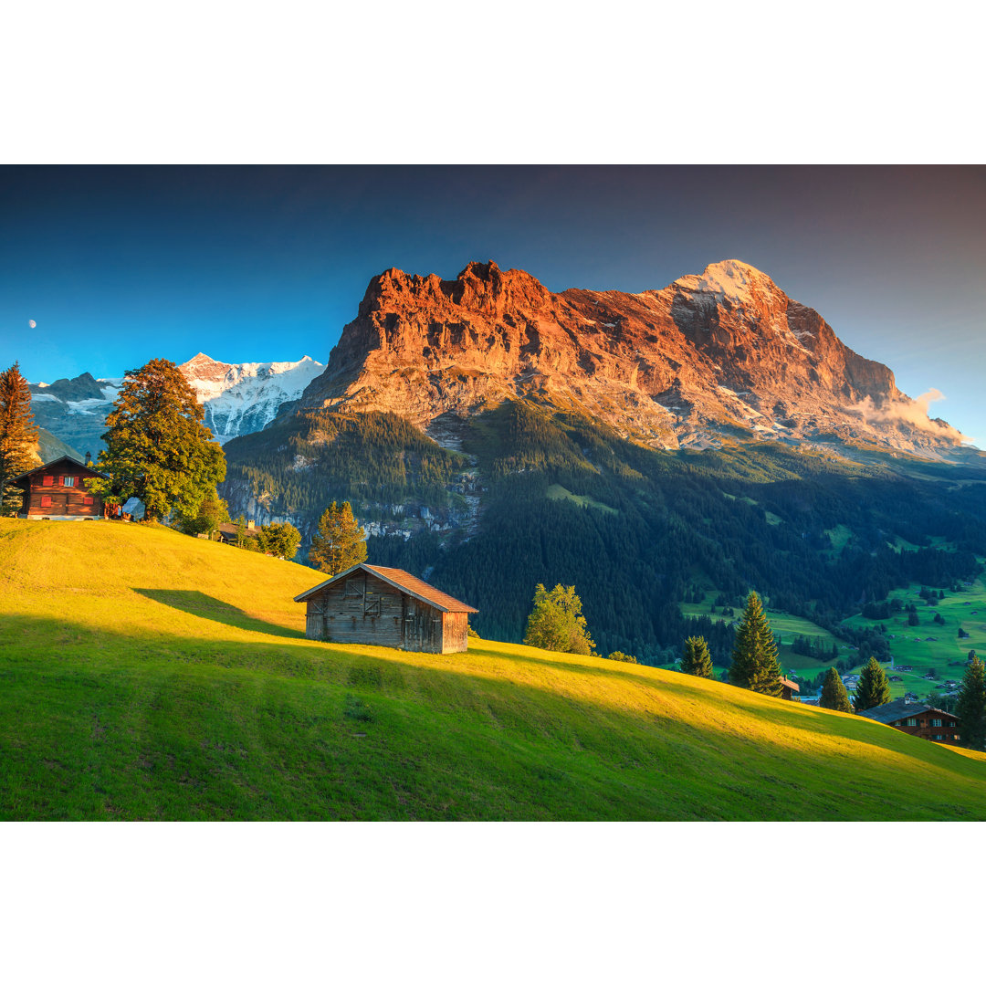 Leinwandbild Alpine Chalets mit grünen Feldern und hohen Bergen bei Sonnenuntergang