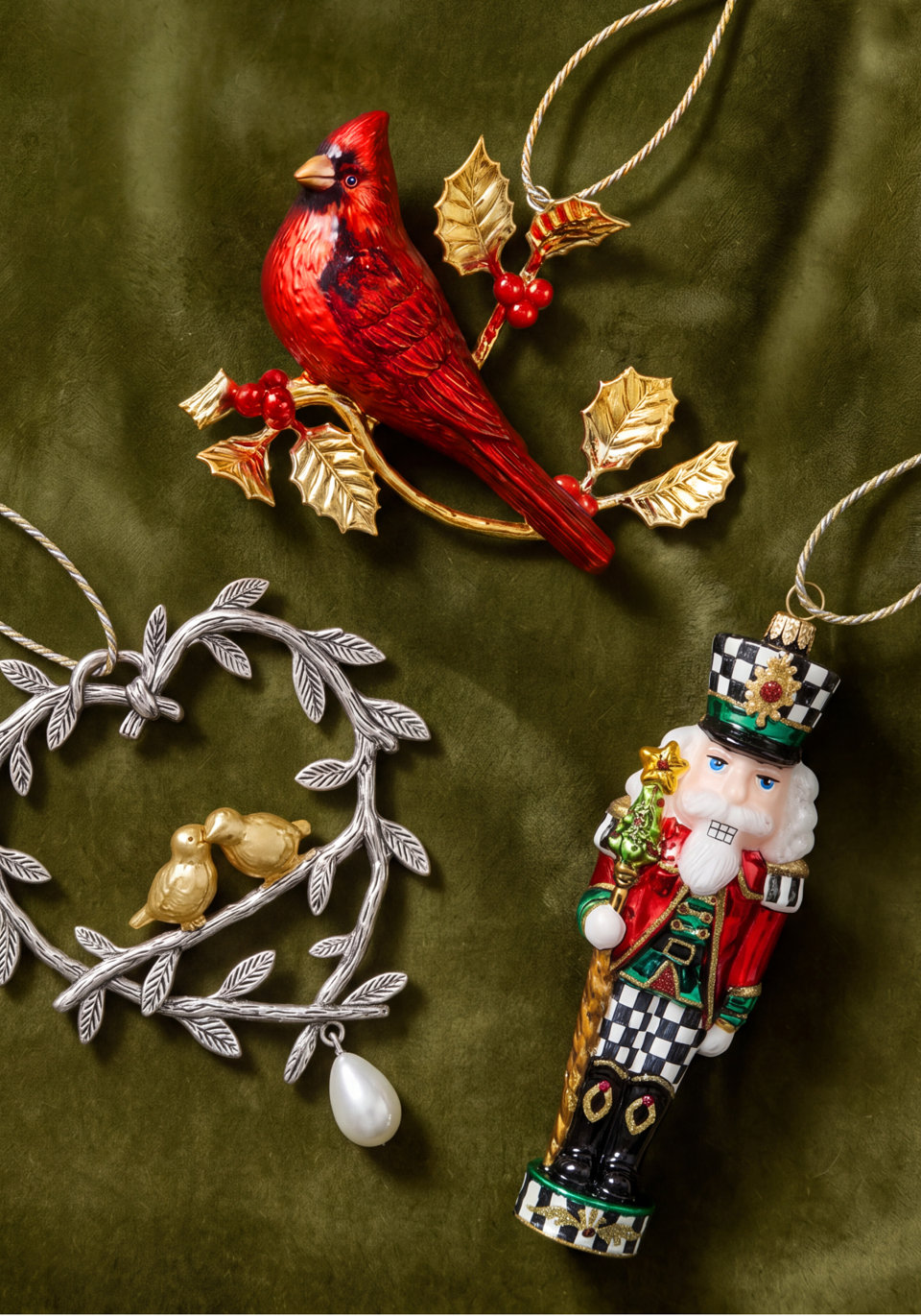 Three decorative Christmas ornaments including a red cardinal perched on a gold holly branch, a silver heart-shaped wreath with two gold birds, and a nutcracker in a black and white checkered uniform, all displayed against a dark green velvet background.