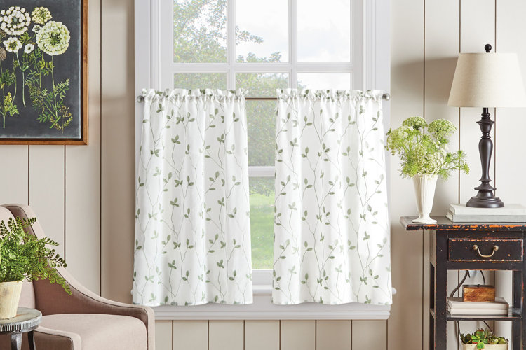 white café curtains with green vine print on sunny window above side table and beige armchair