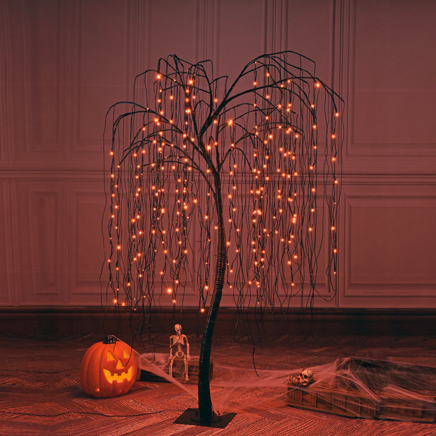 Pre-lit artificial weeping willow tree with orange LED fairy lights, placed on a floor with a jack-o'-lantern, small skeleton, and decorative spiderwebs for Halloween decor.