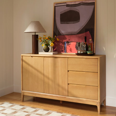 Light oak sideboard with two doors and three drawers, styled with a lamp, flowers, and art against a white paneled wall.