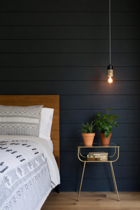 Modern bedroom with a dark wood plank accent wall, a wooden bed frame with patterned bedding, a gold metal nightstand with potted plants, and a hanging pendant light.