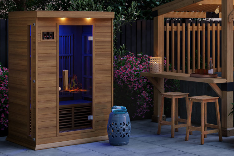 Small wooden sauna placed on a patio beside a blue garden stool and a pergola with a built-in bar on the side.