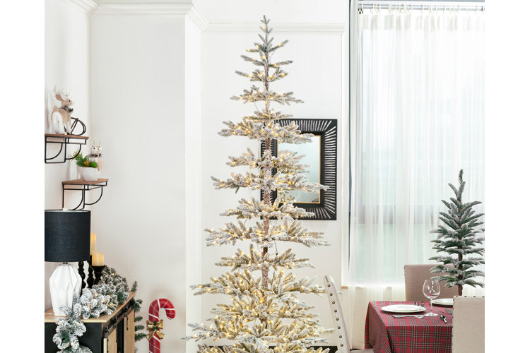 A sparse prelit Christmas tree with flocked branches placed in a corner between a dining room sideboard and a dining table.