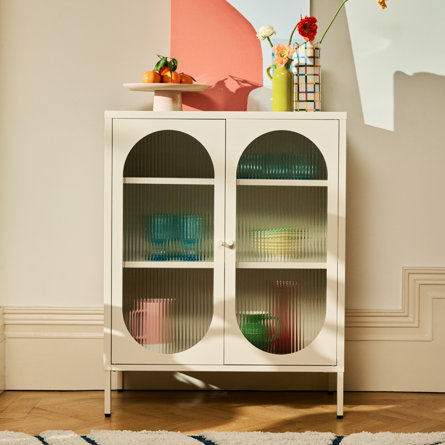 A white cabinet with fluted glass doors displaying colourful items inside, topped with decorative vases and a fruit bowl