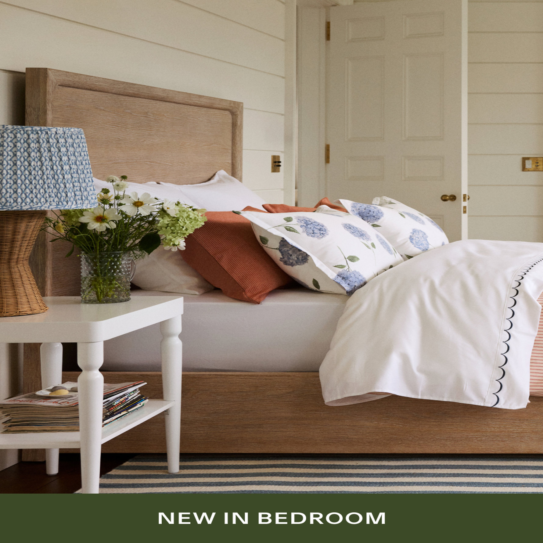 A cozy bedroom featuring an oak bed frame with hydrangea-patterned pillows, rust and white bedding, a white nightstand with fresh flowers, and a wicker lamp with blue patterned shade. Text reads 'NEW IN BEDROOM'.