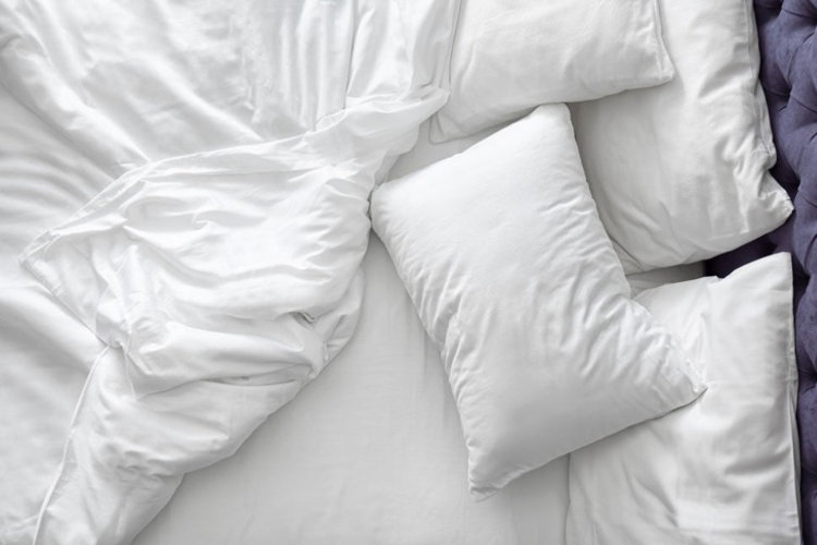 unmade bed with white bedding and layered pillows in front of a purple upholstered headboard