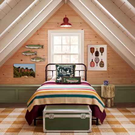 Chambre à coucher au style rustique avec plafond mansardé en bois, lit en métal habillé d'une couverture rayée, décorations murales sur le thème de la pêche et malle verte au pied du lit.