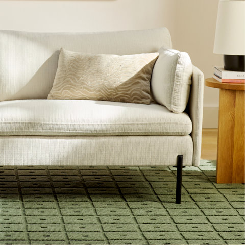 Cream sofa with a beige textured lumbar pillow sitting on a sage green grid-patterned rug next to a small wooden side table and lamp.