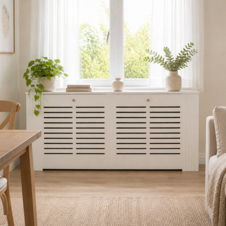 White wooden radiator cover with horizontal slats positioned under a bright window, styled with potted plants and decorative items in a living room setting.