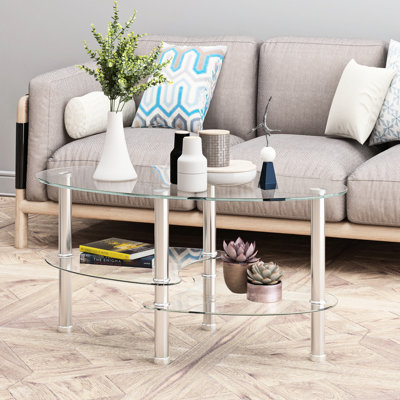 Table basse moderne en verre transparent avec pattes en acier inoxydable, de forme ovale et à trois niveaux, idéale pour le salon. Meuble polyvalent et fonctionnel.