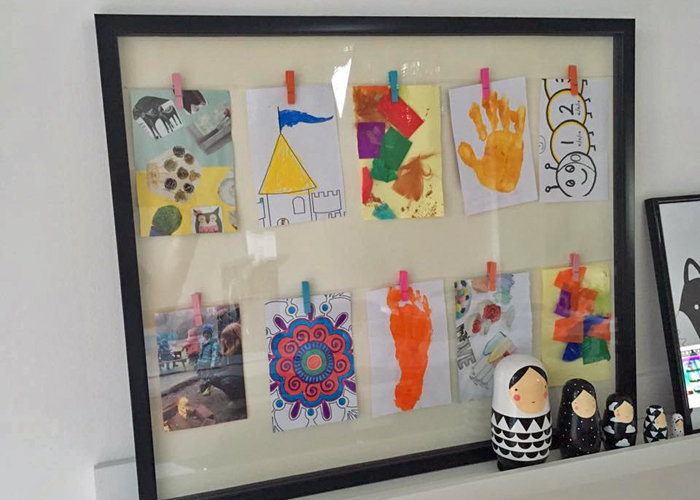 A large black picture frame with white backing and clotheslines displaying children's artwork on ten pegs