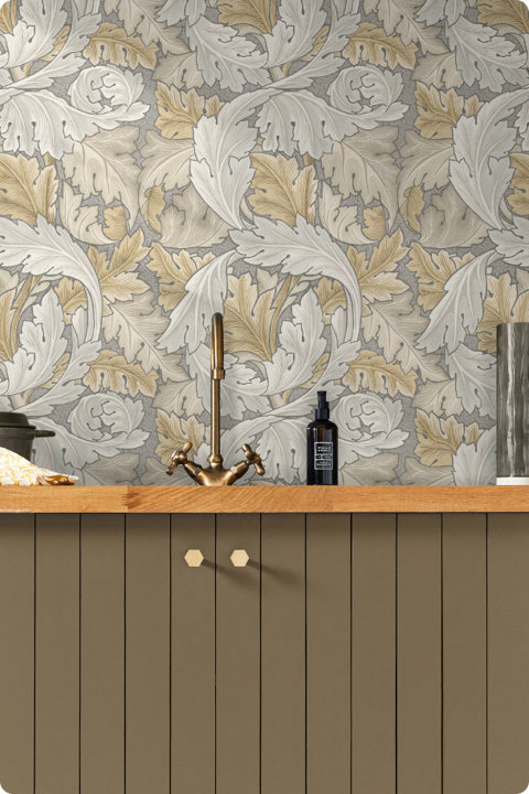 A kitchen backsplash featuring William Morris style ornate acanthus leaf wallpaper in shades of cream, gold, and grey, situated behind a brass faucet and a wooden countertop with olive green cabinetry.