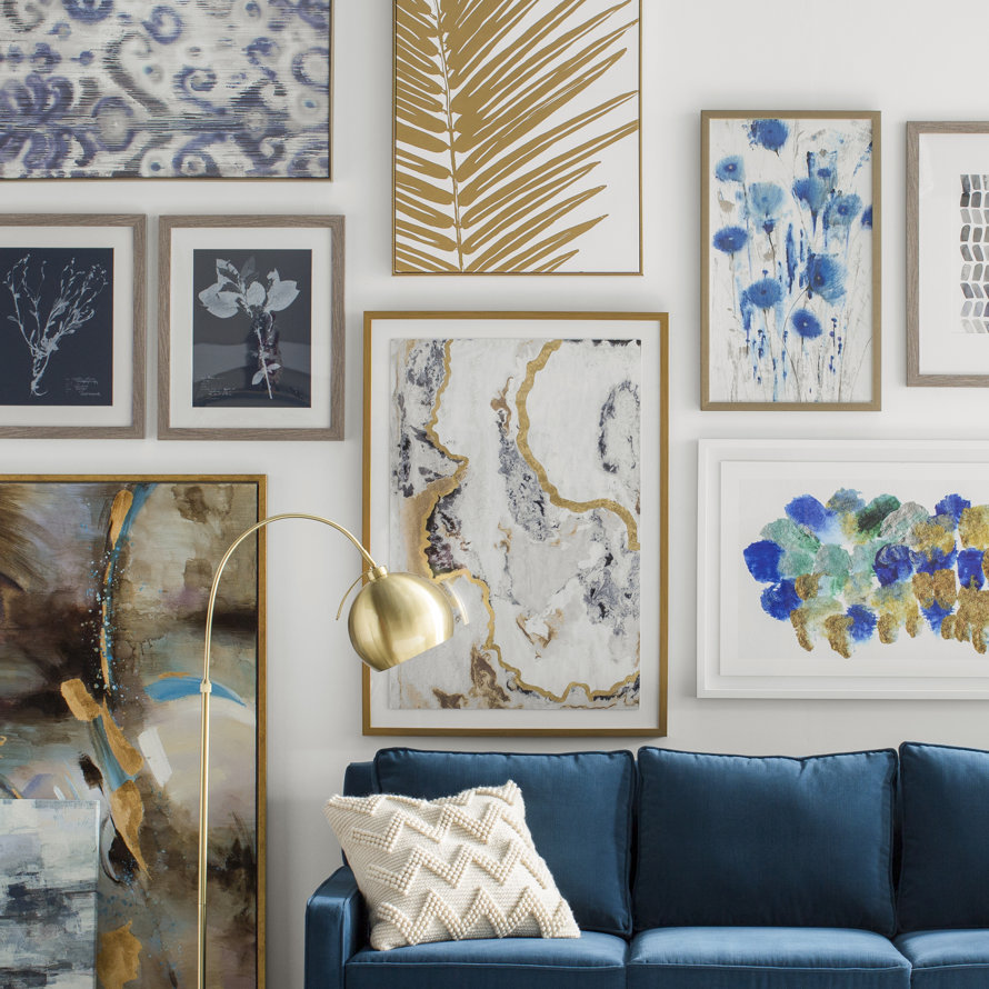 Gallery wall featuring a collection of framed abstract, floral, and botanical artwork in gold, blue, and white tones hung above a deep blue velvet sofa with a textured cream pillow and a gold floor lamp.