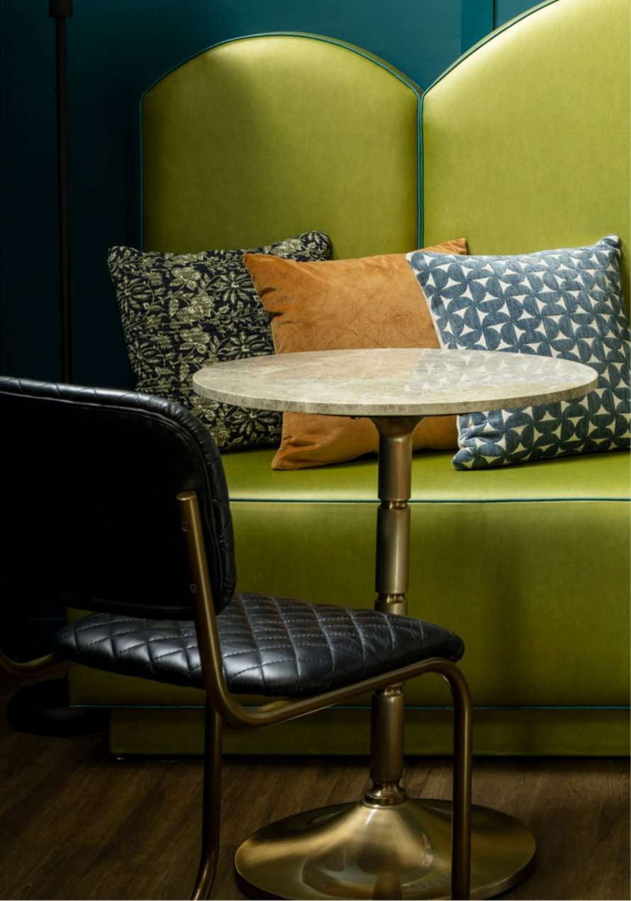 Upholstered booth seating with decorative pillows and a small marble table next to a black, cushioned chair.