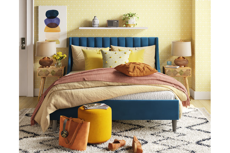 Blue upholstered bed in a yellow wallpapered room. Yellow duvet with matching bed and throw pillows and a peach throw blanket. White geometric shag area rug with a yellow velvet ottoman. Two wood mid-century modern nightstands with two gold and white table lamps.