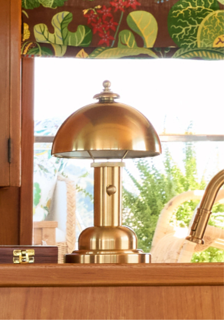 A polished brass mushroom-style table lamp sits on a wooden surface in front of a window with botanical print valances.