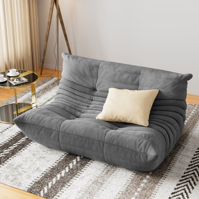 Large grey suede bean bag sofa with tufted detailing and a cream throw pillow in a modern living room.