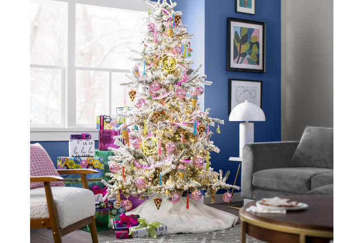 Flocked tree with pink baubles, pizza decor, and other cute tree decorations. White fur tree skirt. Two accent chairs and a stack of wrapped Christmas gifts behind the tree