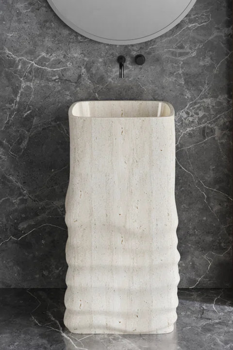 Freestanding beige travertine pedestal sink with a wavy textured base against a dark gray marble wall.