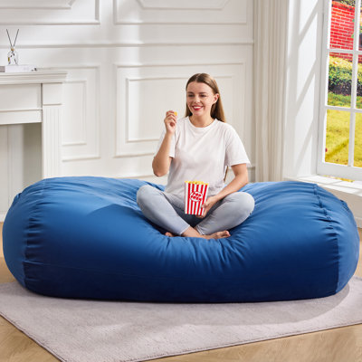 Pouf géant de 1,80 m en velours bleu, sans os et en mousse mémoire compressée. Housse amovible et poche latérale pour salon ou chambre.
