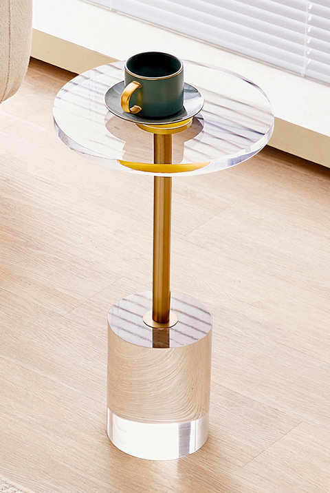 Modern round side table featuring a clear acrylic top and base connected by a gold metal pedestal, with a small mug placed on top.