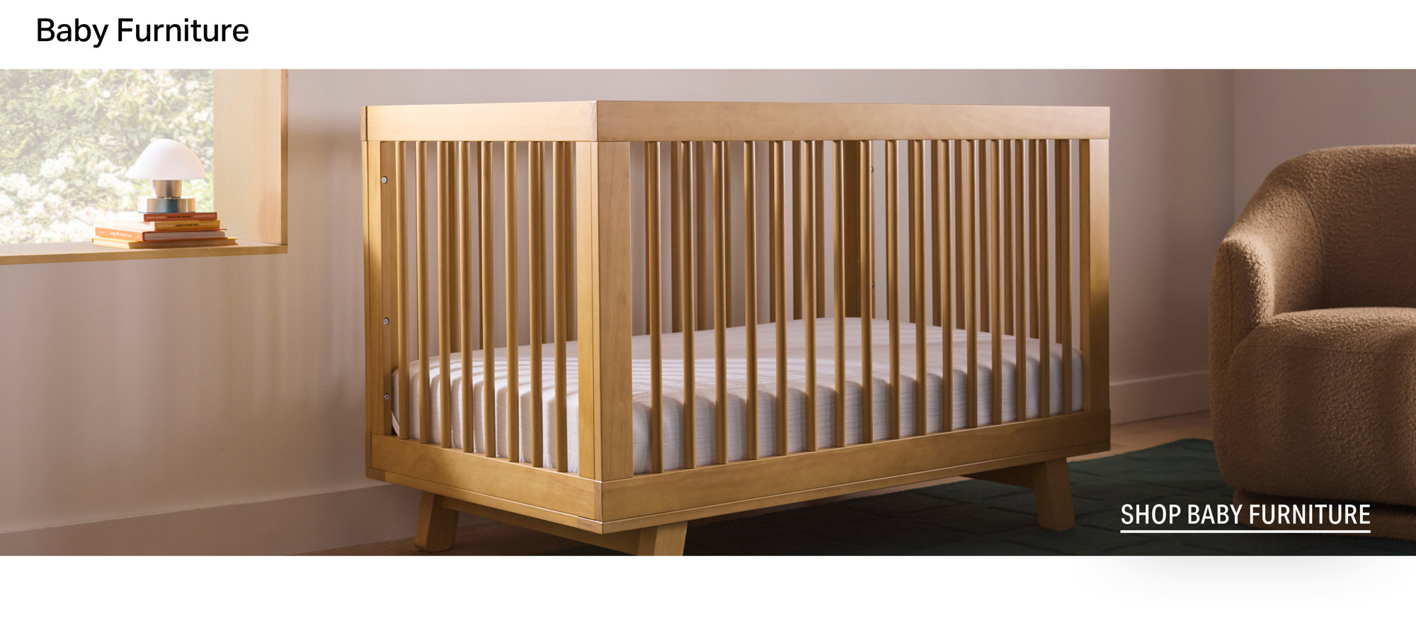Light wood crib in a nursery with framed wall art, a colorful mobile, and a green textured rug, with the text Shop Baby Furniture.