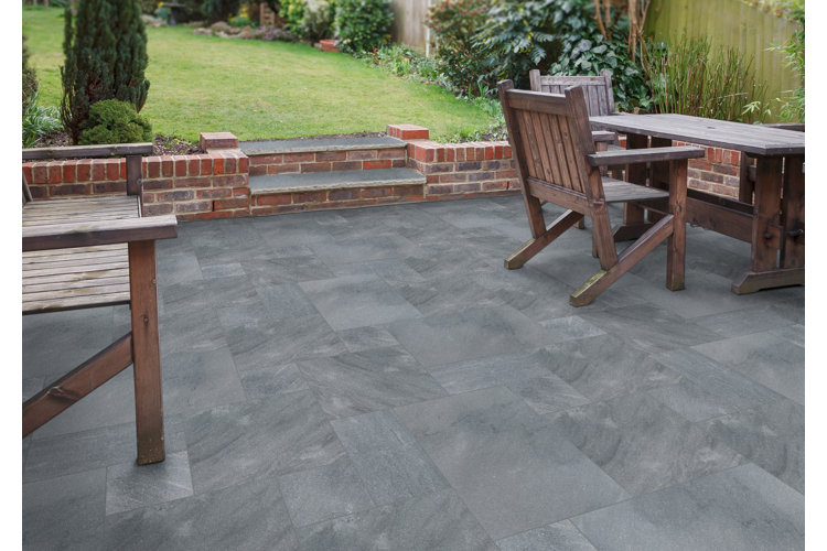 Backyard patio with gray paving stones, wood furniture and a brick border separating the patio from a grass lawn.
