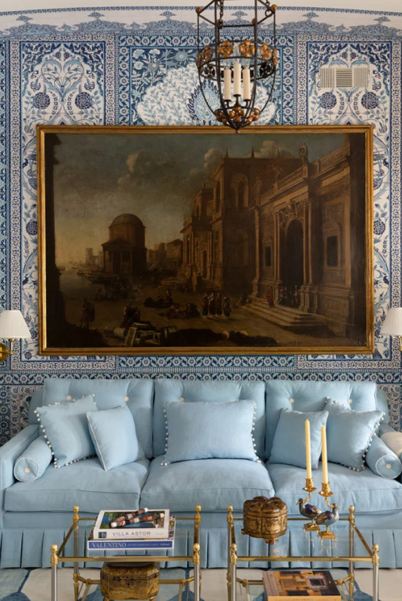 Light blue couch with throw pillows in a living room with blue wallpaper and a historic painting.