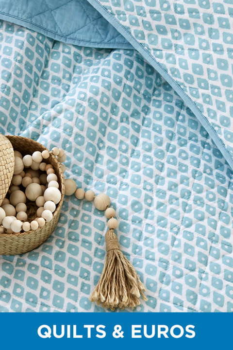 Close-up of a blue and white geometric patterned quilt with a decorative bowl of wooden beads and tassel, featuring the text Quilts & Euros.