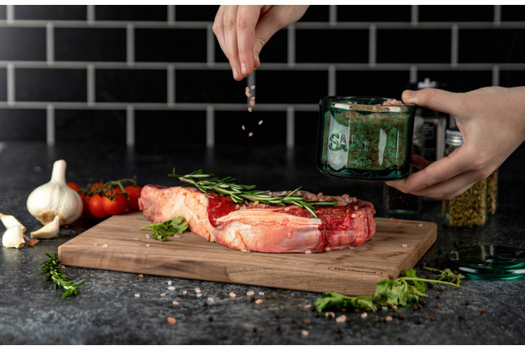 Hands holding a green glass salt cellar and sprinkling coarse salt on meat